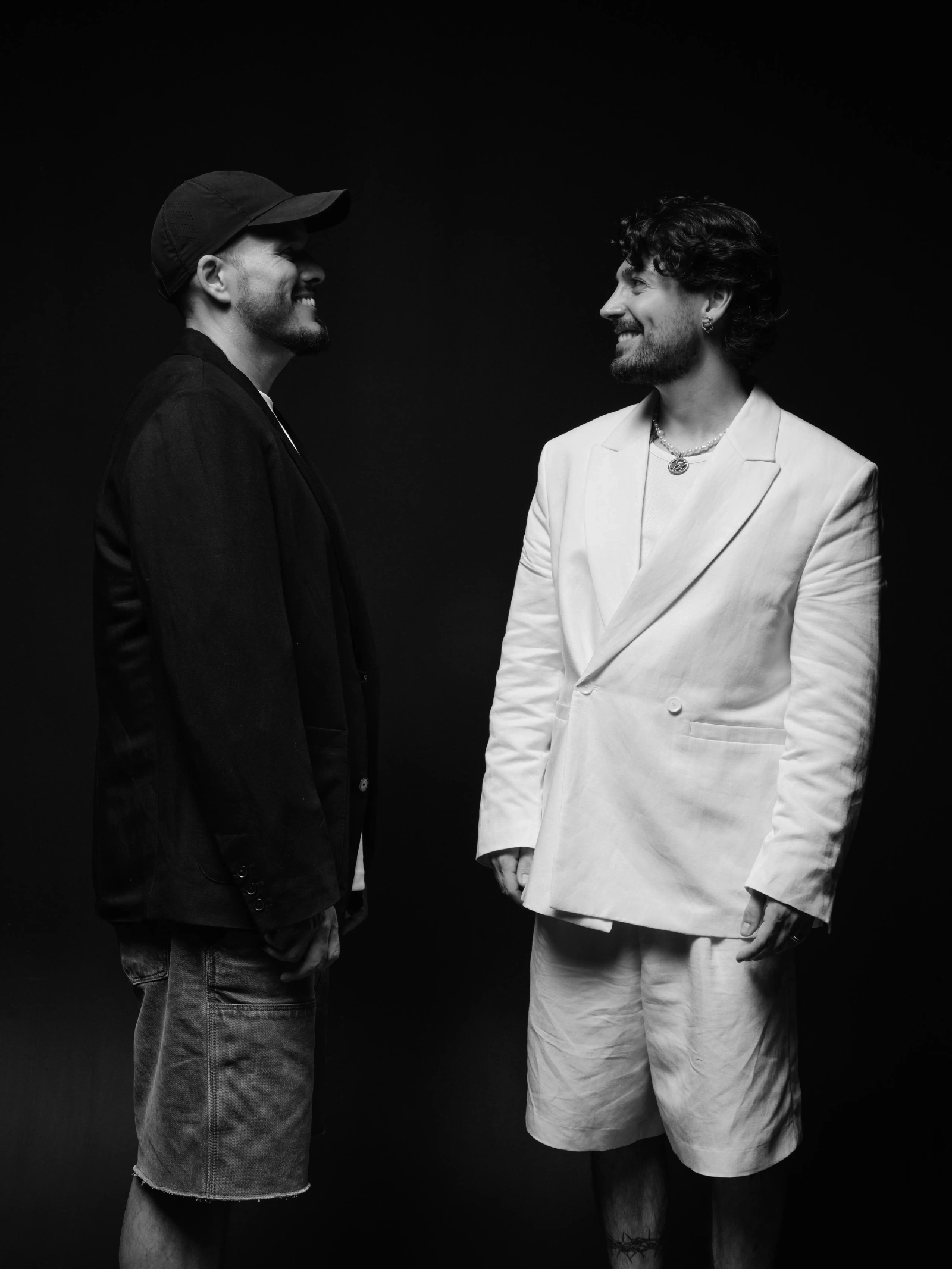 two men facing each other and smiling, one in a dark jacket and cap, one in a white suit, black background
