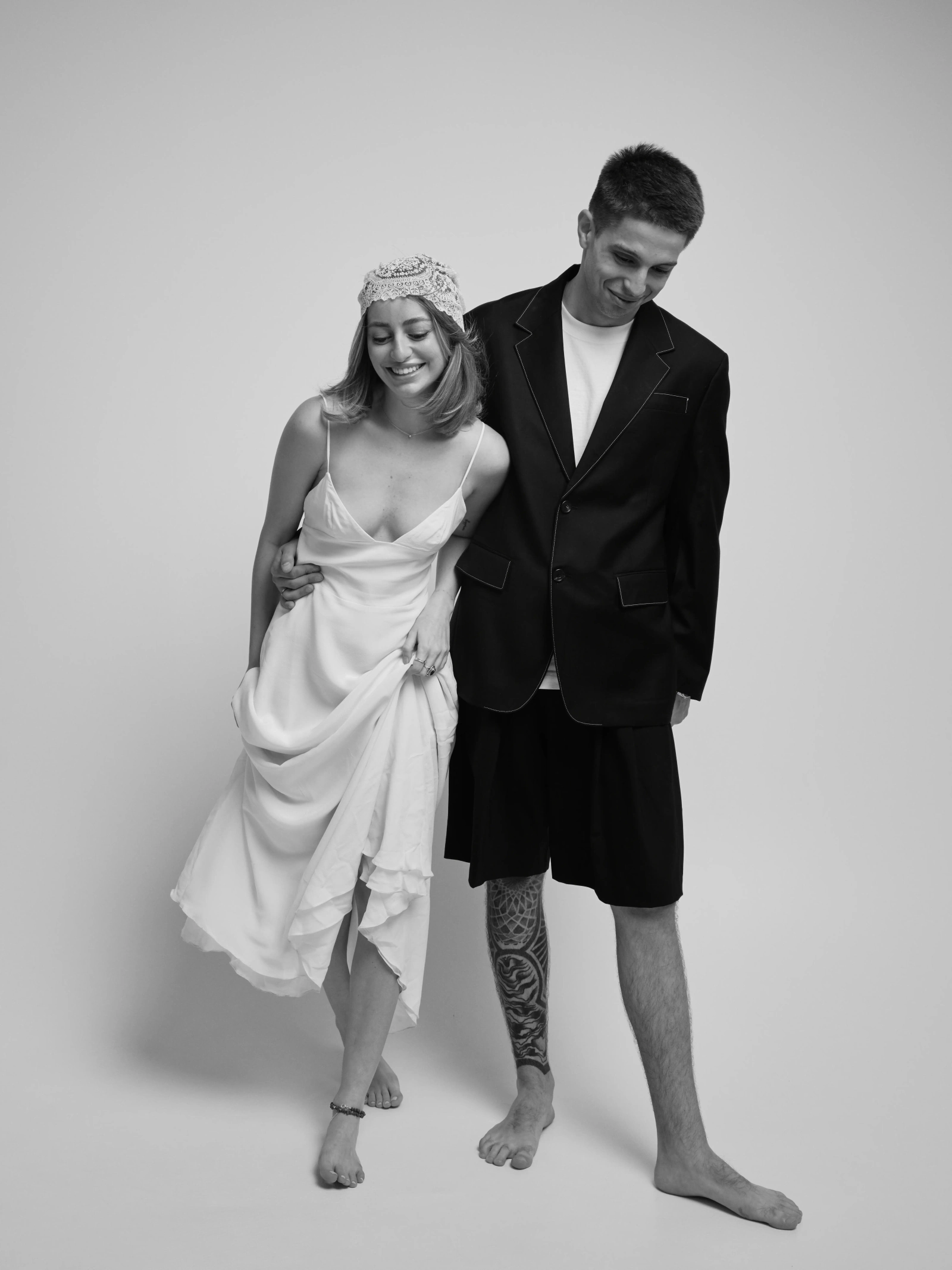 a couple walking barefoot, she in a white dress, he in a dark blazer and shorts, white background
