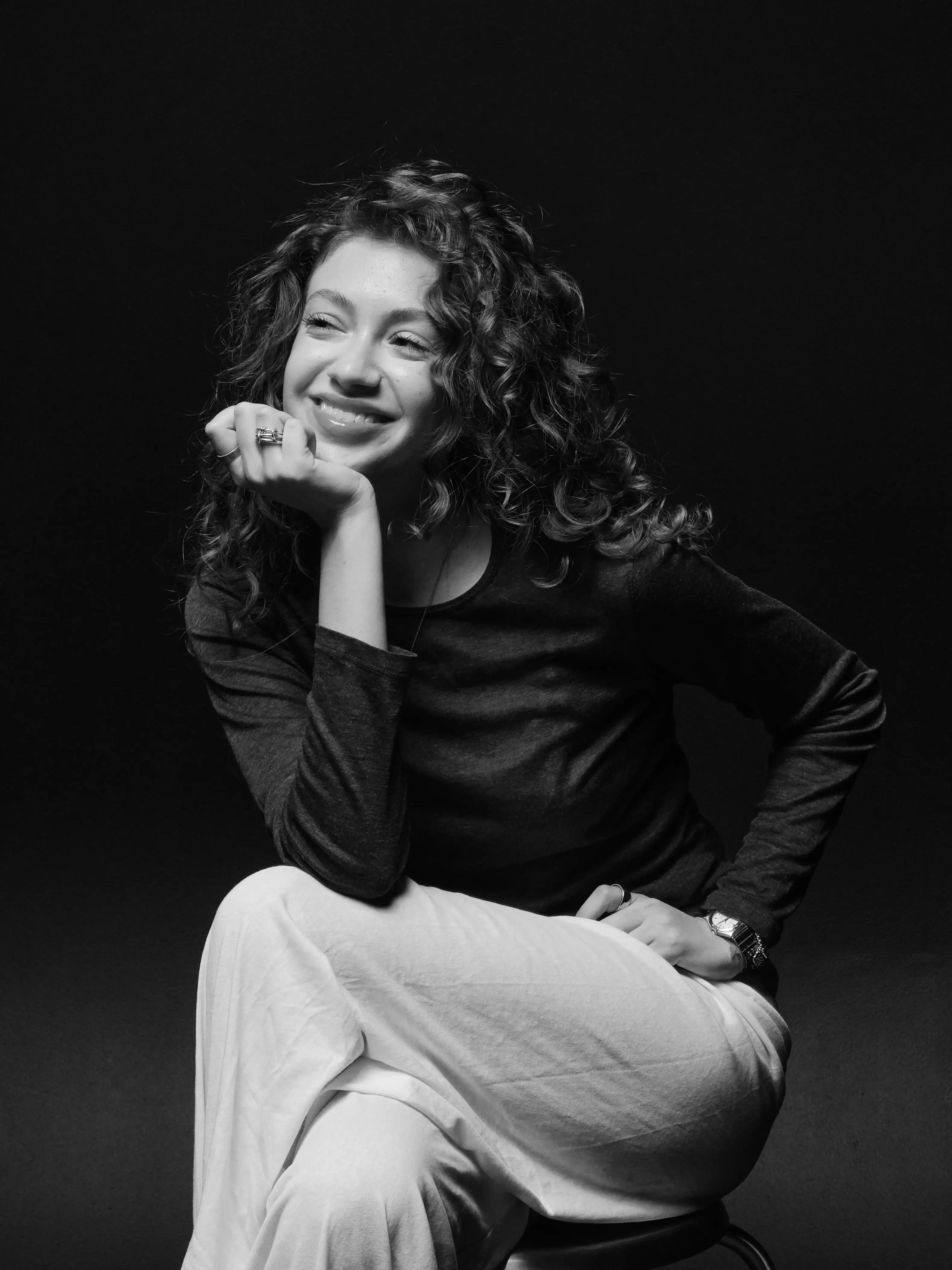 a woman with curly hair smiling, chin resting on her hand, black background