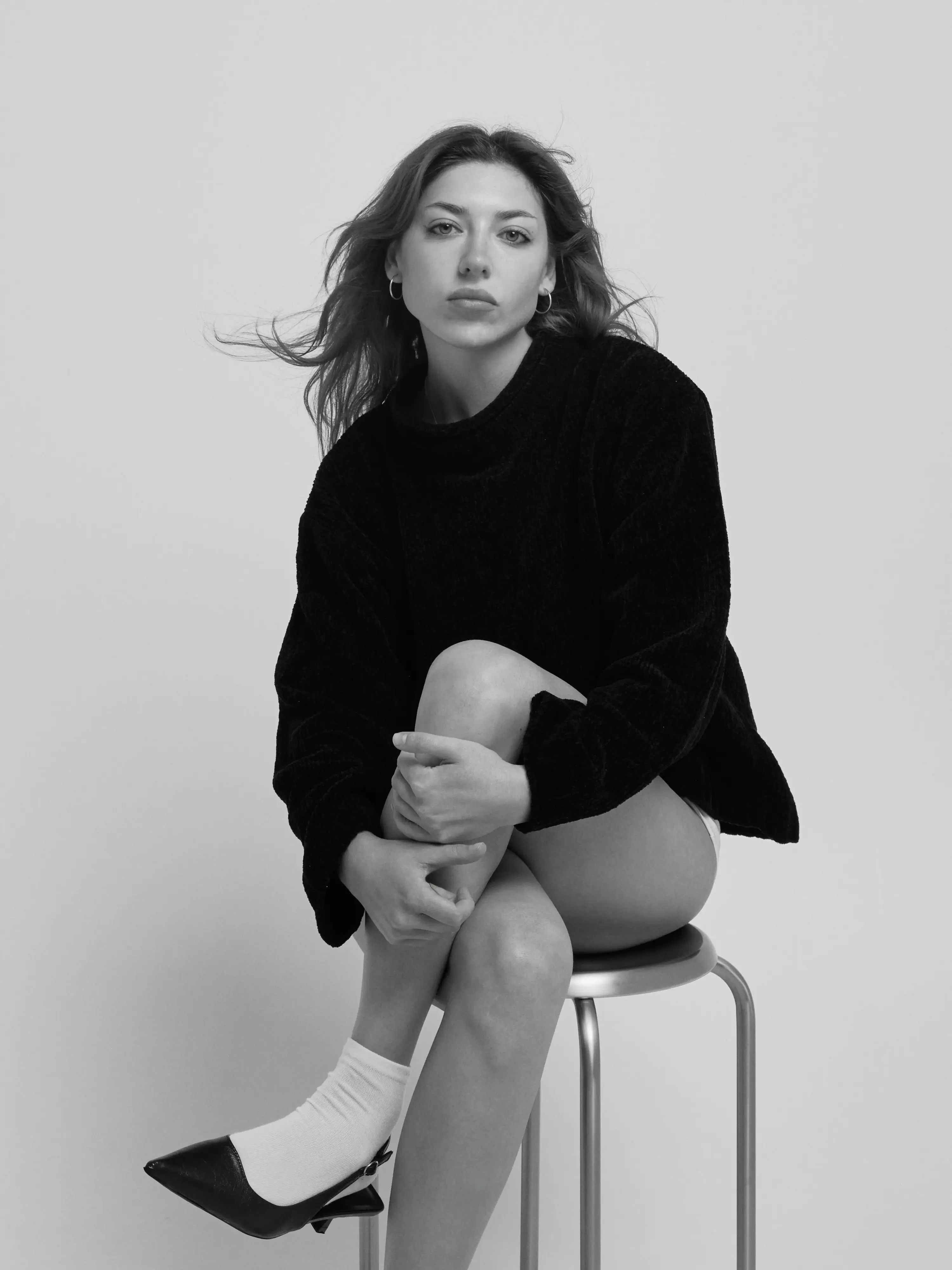 a woman sitting on a stool hugging one knee, her hair moving, white background