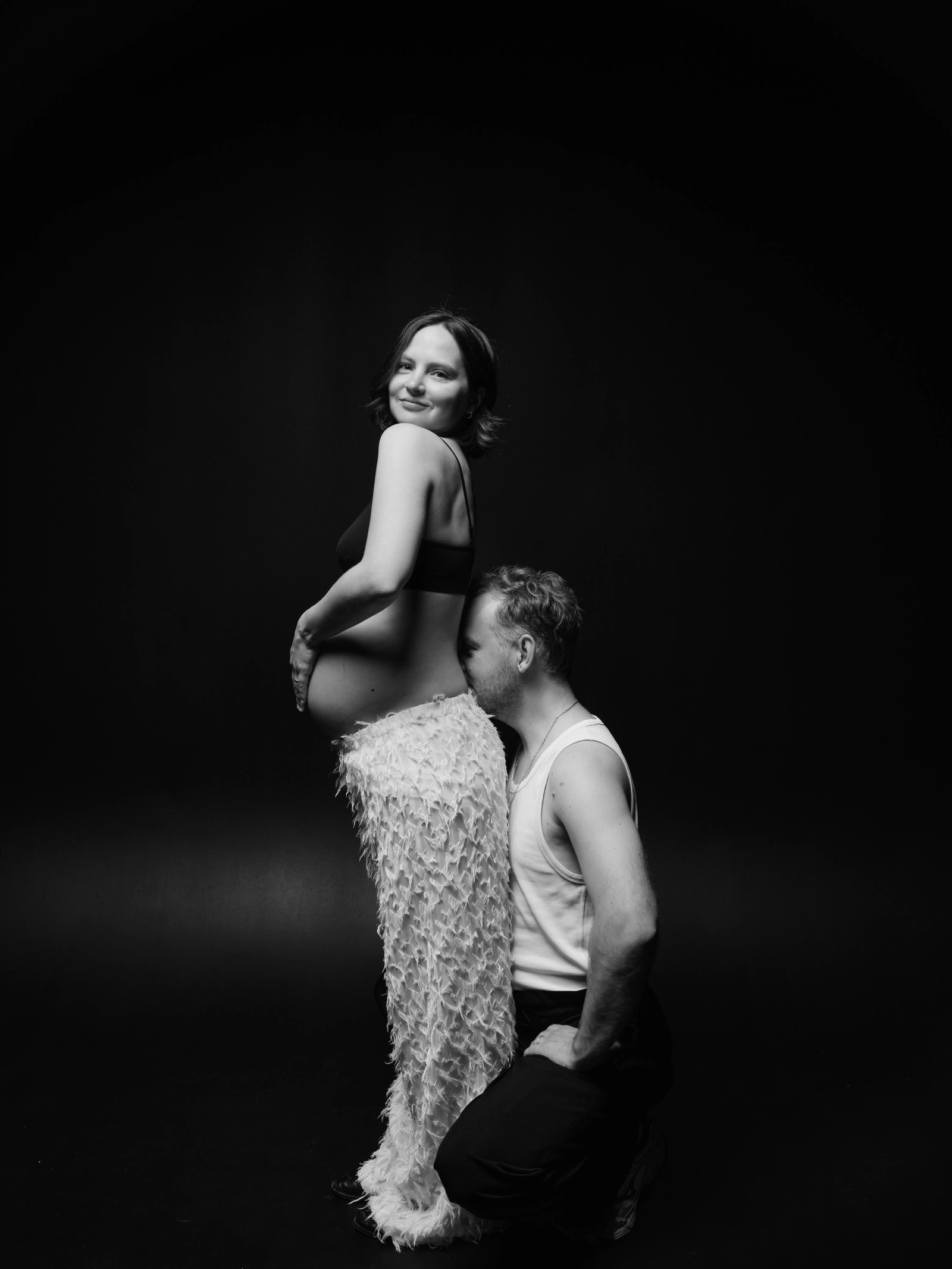 a pregnant woman standing while her partner kneels with his head against her belly, black background