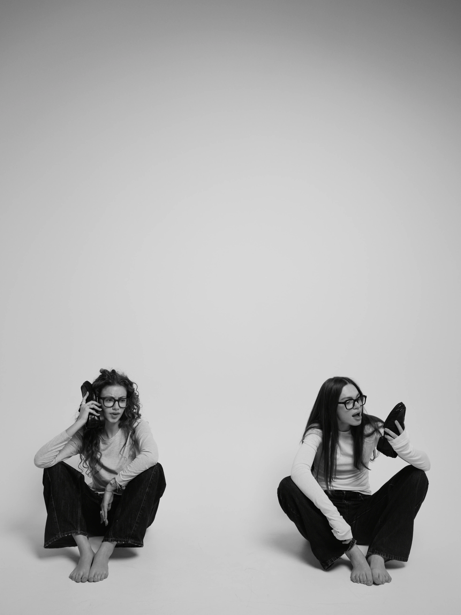 two friends sitting on the floor, each holding a shoe to her ear like a phone, white background